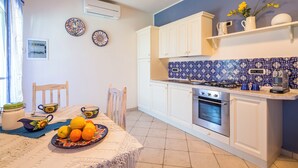 Una cocina compacta con gabinetes blancos, una estufa y un lavamanos. Hay platos decorativos en la pared y una mesa con frutas y una tetera.