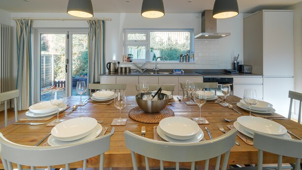 Un coin repas avec une table en bois dressée pour six personnes, des chaises blanches et des luminaires suspendus.