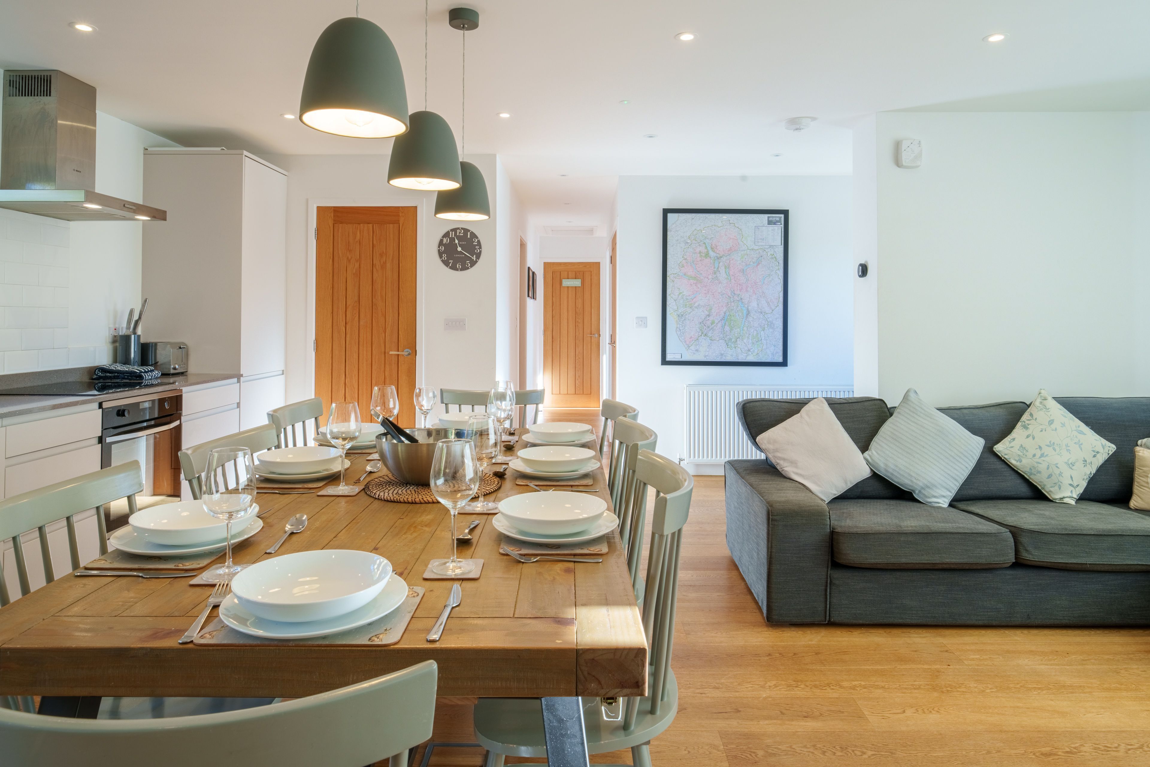 A dining area with a wooden table set for a meal, a grey sofa, and a kitchen in the background.