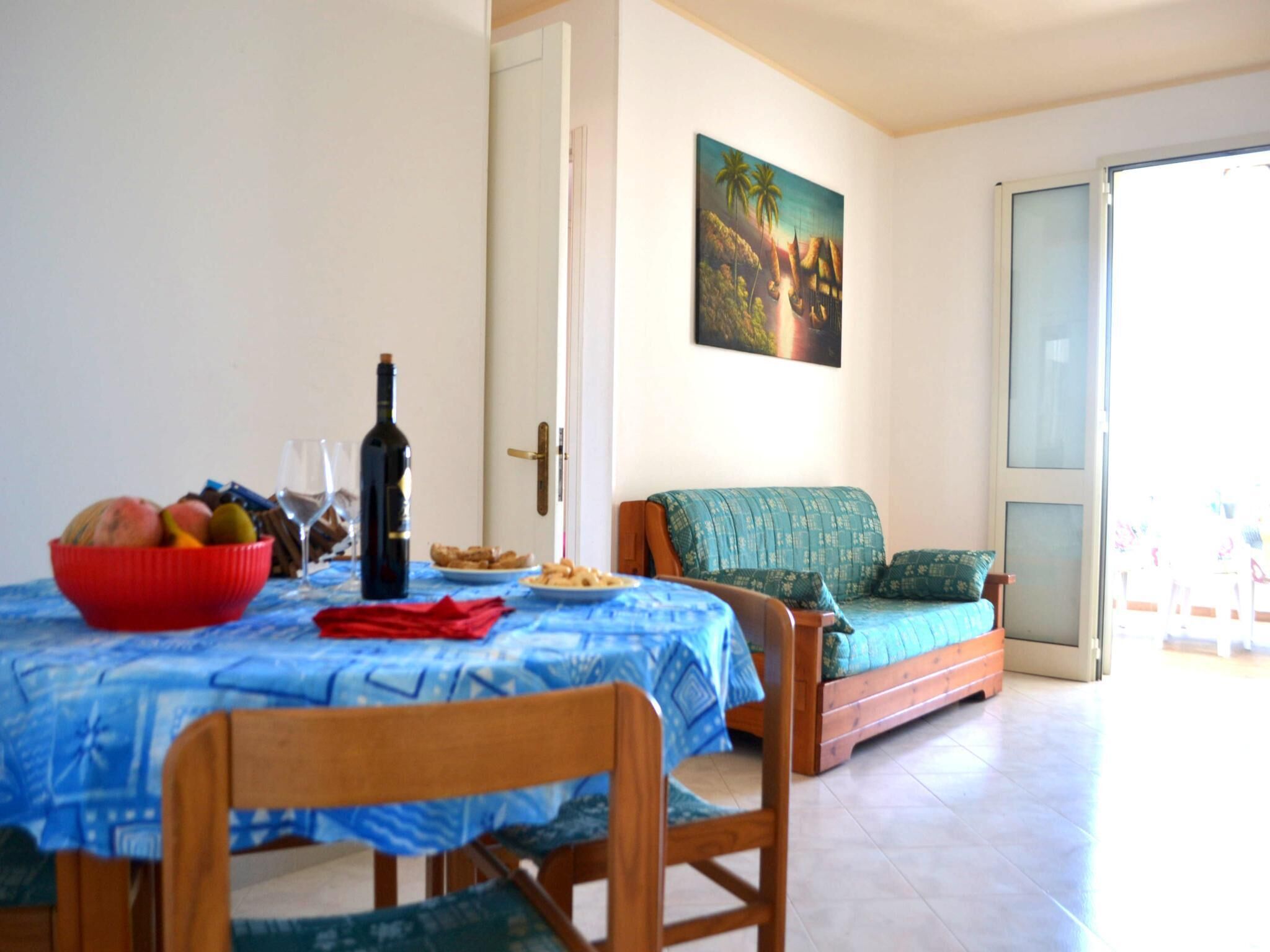 A dining area with a table set for two, a bowl of fruit, a bottle of wine, and a plate of snacks. There is a sofa with a patterned cushion and a painting on the wall.