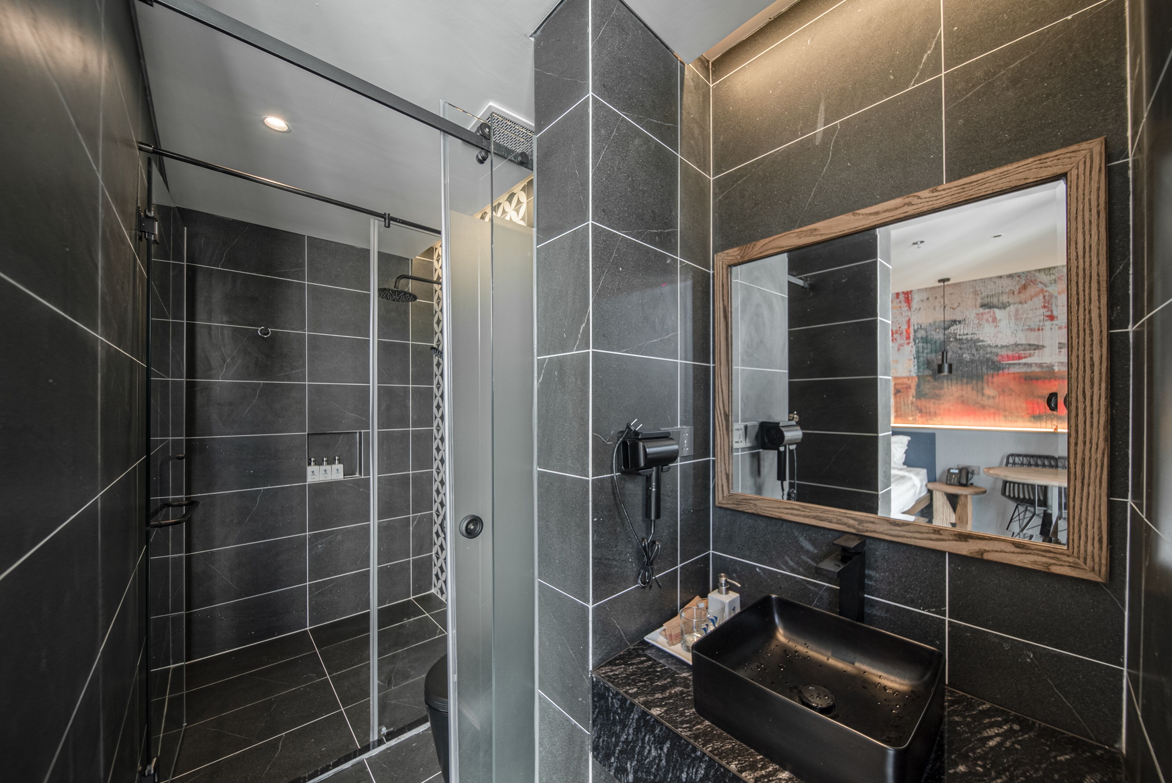 A modern bathroom with dark tiles, a glass shower enclosure, a sink with a wooden frame mirror, and a towel rack.
