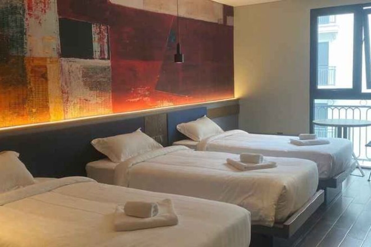 Habitación de hotel con tres camas, un cuadro abstracto grande en la pared y vistas de la ciudad a través de una ventana.