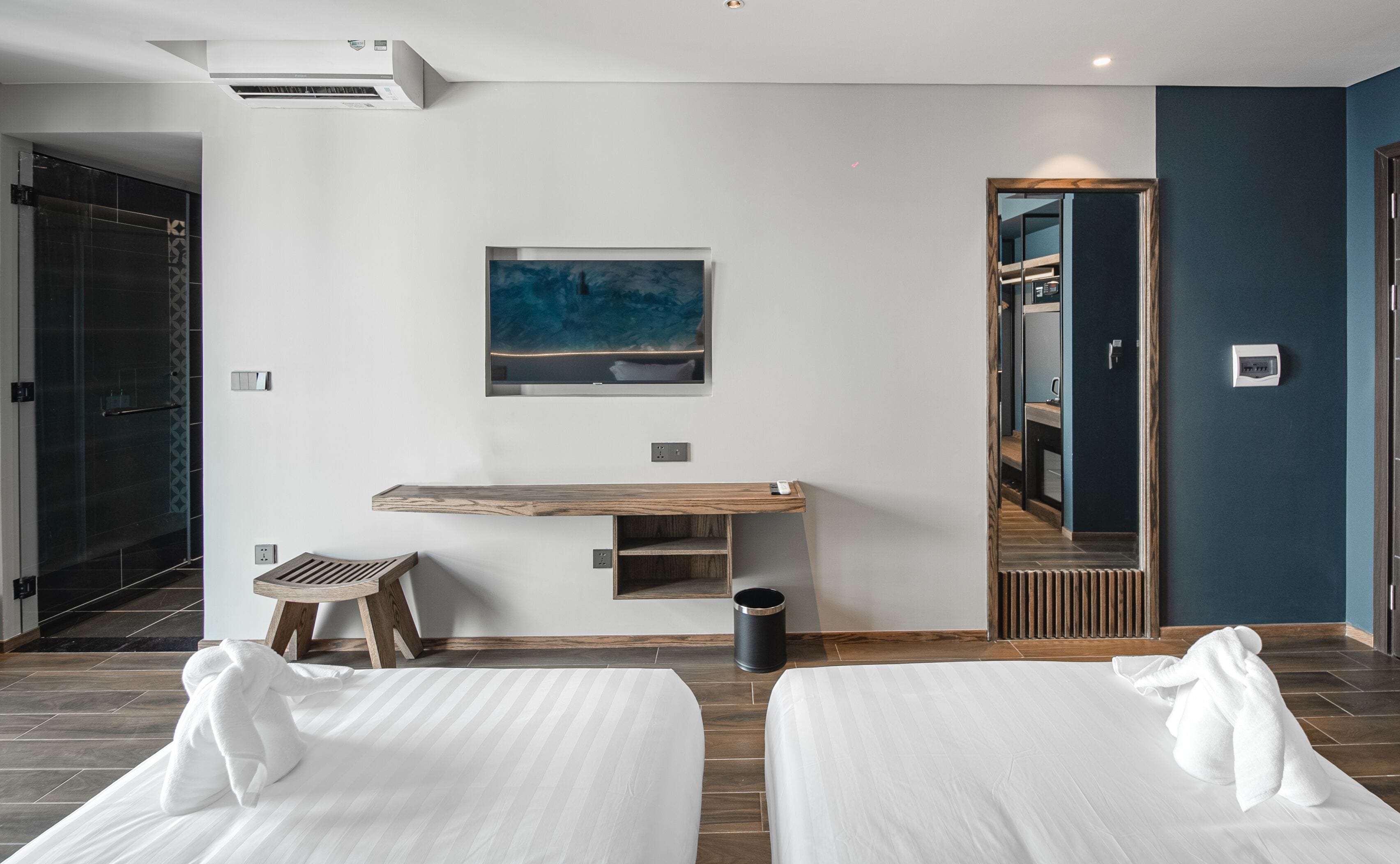 A modern hotel room with two beds, a wooden bench, a painting, a mirror, and a shower area.