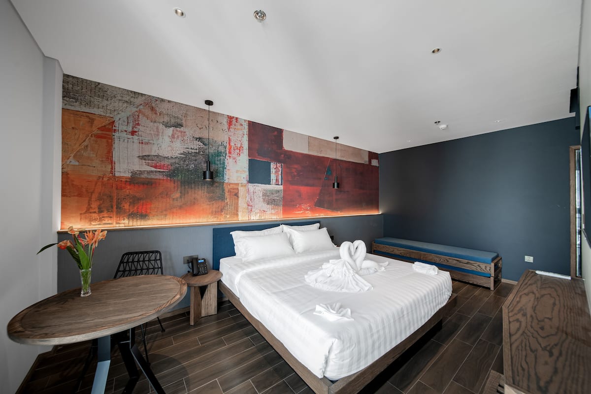 Una habitación de hotel moderna con una cama grande, una mesita con un florero, un banco y un cuadro abstracto de gran tamaño en la pared.