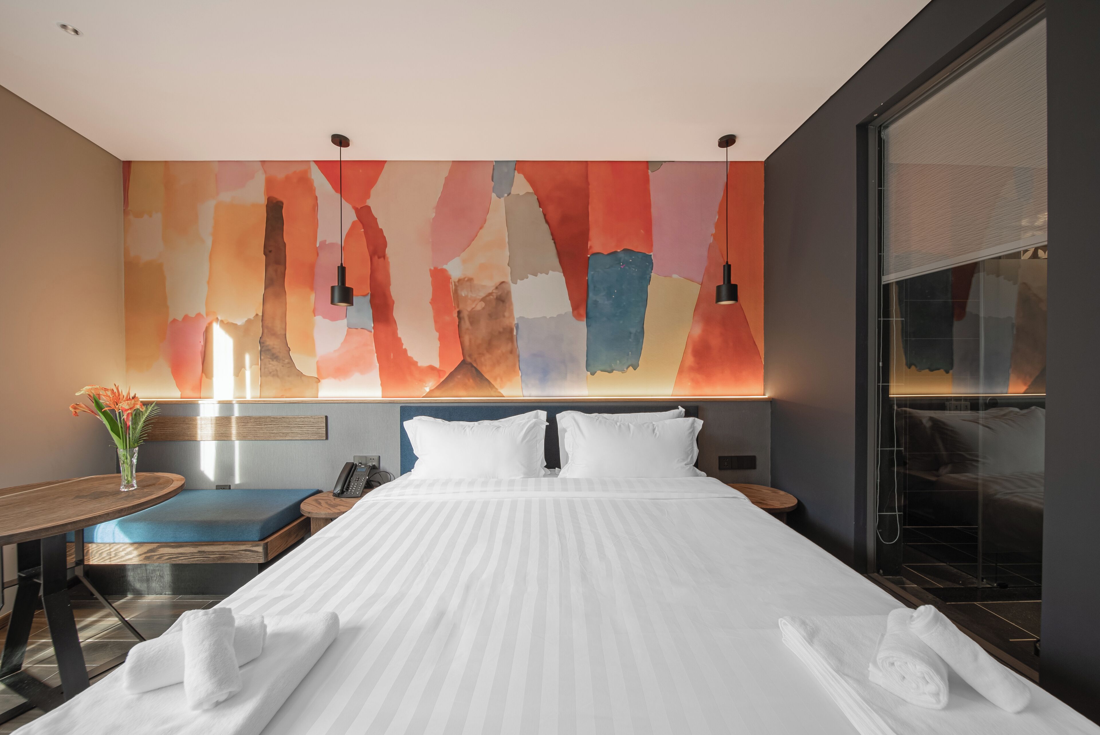 Une chambre d’hôtel moderne avec un grand lit, une table de chevet avec une lampe, un petit coin salon et un grand tableau abstrait accroché au mur.