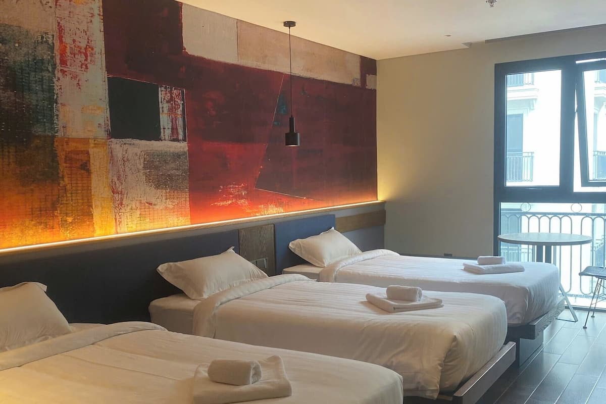 Habitación de hotel con tres camas, un cuadro abstracto grande en la pared y un balcón con vistas a la ciudad.