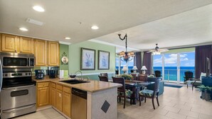 Una cocina con armarios de madera, electrodomésticos de acero inoxidable y una isla grande. Hay un comedor con mesa y sillas, y vista al océano a través de amplios ventanales.
