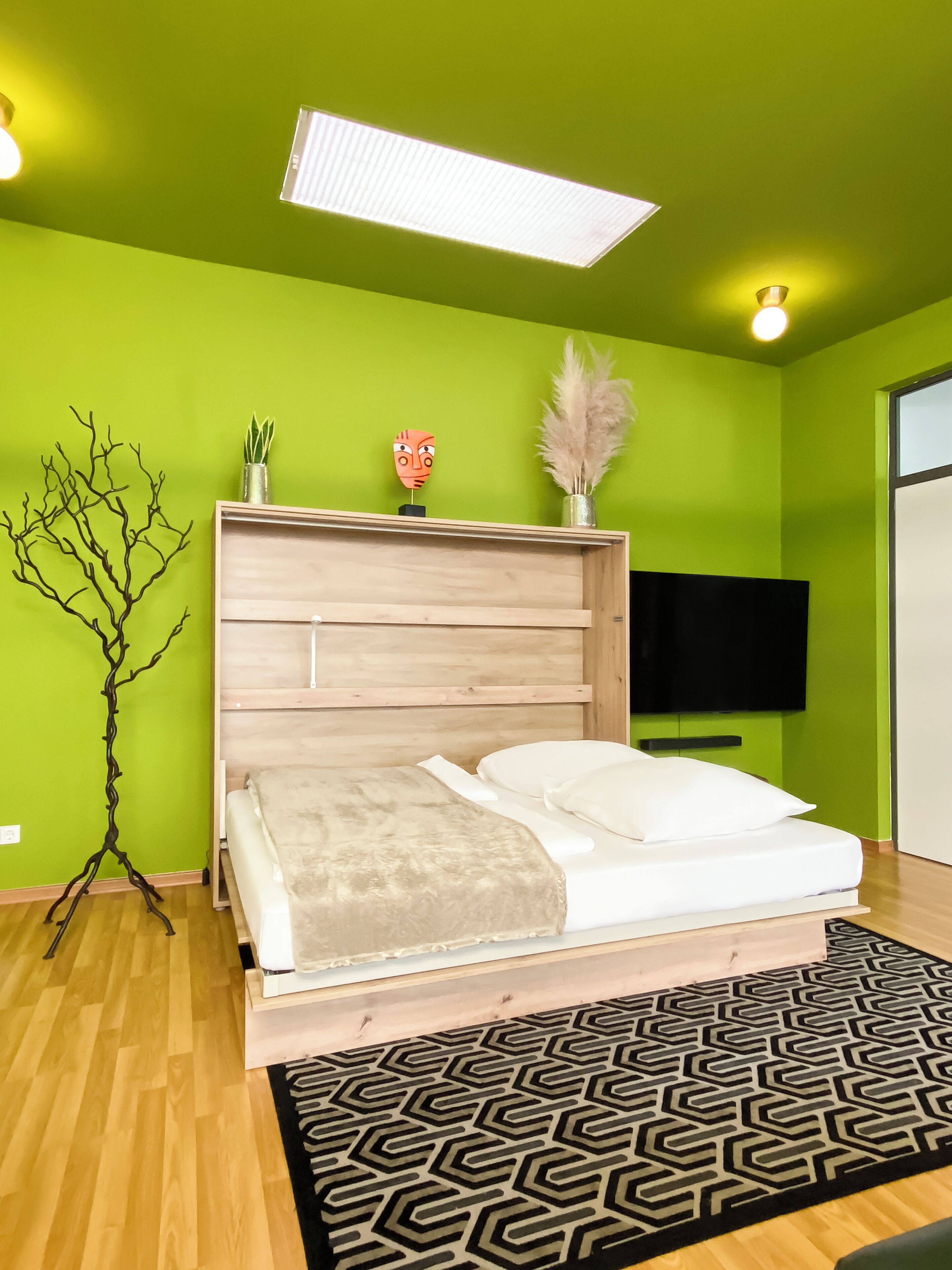 Ein Schlafzimmer mit einem hölzernen Bett, grünen Wänden, einem schwarz-weißen Teppich, einem an der Wand montierten Fernseher und einer Deckenleuchte.