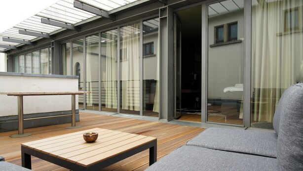 A modern outdoor patio with a wooden table and grey sofa, large glass doors leading to a room, and a metal pergola overhead. - Exklusives City Apartment Nature (Leipzig)