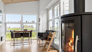 Un intérieur moderne avec une grande cheminée, un coin repas avec des chaises et de grandes fenêtres donnant sur l’extérieur.