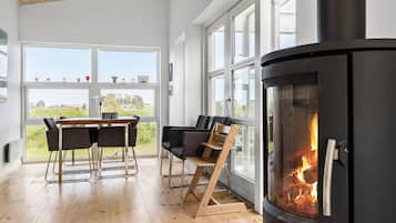 Et moderne interiør med en stor pejs, et spiseområde med stole og store vinduer med udsigt til omgivelserne.