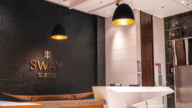 A reception area with a dark textured wall, a wooden reception desk, and two pendant lights. - Sway Suites (Manama)