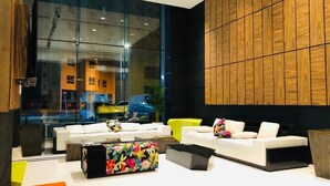A modern lobby with a glass wall, white sofas, a colorful floral bag, and a wooden panel wall. - Sway Suites (Manama)