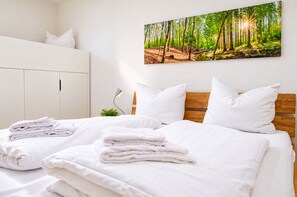 A neatly made bed with white linens and pillows, a white wardrobe, and a wall-mounted painting of a forest.
