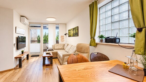 A modern living room with a sofa, a coffee table, and a dining table. There is a window with curtains, a flat-screen TV mounted on the wall, and a small plant on the windowsill.