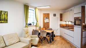 Ein kleiner Wohnbereich mit einer Couch, einem Esstisch, Stühlen, einer Küchenzeile und einem Fenster mit Vorhängen.