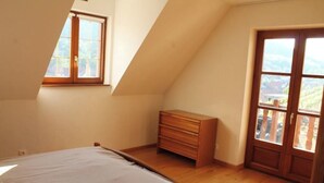 A bedroom with a sloped ceiling, a bed, a wooden dresser, and a large window with a view of greenery. - Maison Située Dans le Vignoble, Appartement à L'étage Avec Entrée Indépendante (Soultzmatt)