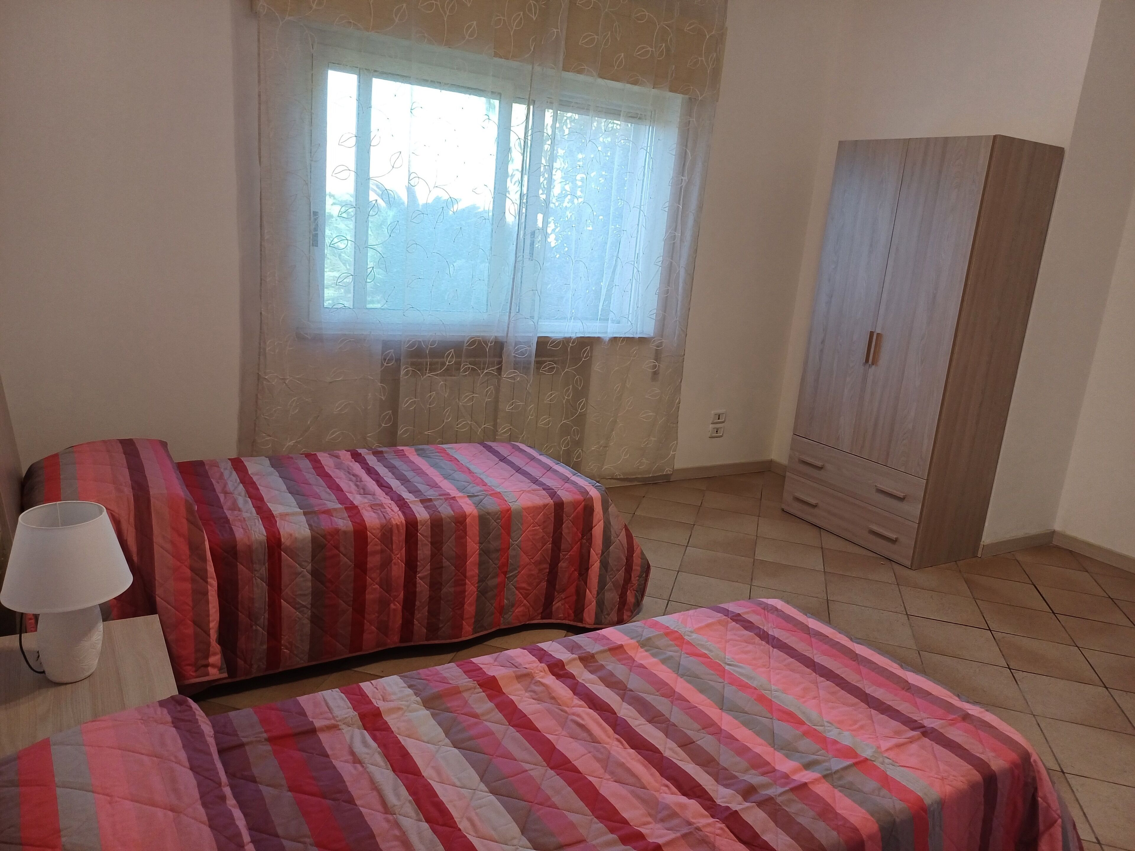 Une chambre avec deux lits, une fenêtre avec des rideaux légers, une armoire en bois et une lampe de chevet.