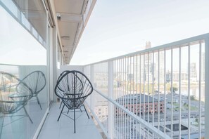 Un balcón con dos sillas de alambre negro y barandilla de vidrio que da a una vista urbana con edificios y calles.