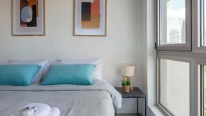 Habitación de hotel con cama, dos almohadas, mesita de noche con lámpara y dos cuadros abstractos en la pared.