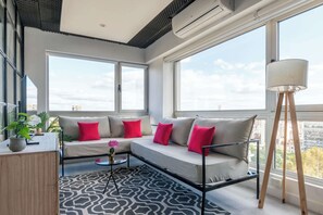 Uma sala de estar moderna com sofá, mesa de centro e abajur. Há janelas amplas que oferecem vista da cidade.