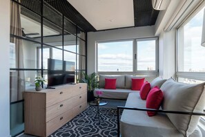 A modern living room with a sofa, TV, and a large window offering a city view.