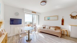 A modern living room with a sofa, a dining table with chairs, a TV mounted on the wall, and a large window offering a view of the sea.