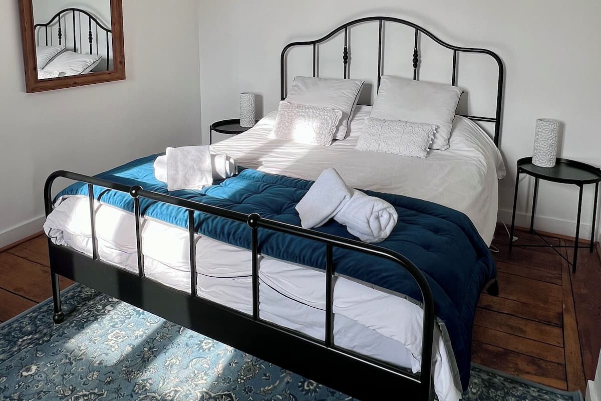 Una cama bien hecha con ropa de cama blanca y azul, un marco de metal negro y un tapete azul estampada. Hay tres cuadros en la pared sobre la cama.