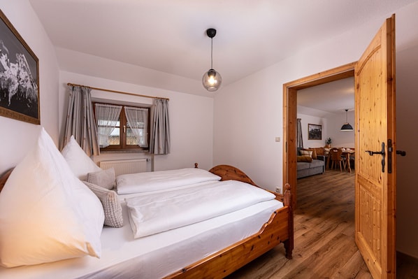 A bedroom with a wooden bed, a window with curtains, a hanging light fixture, and a wooden door leading to a dining area. - Ferienwohnung Scheffmannlehen Nr. 3 (Ramsau bei Berchtesgaden)
