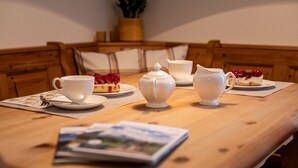 Una mesa de madera con tazas de té blancas, un frutero y una tetera. Hay dos porciones de pastel en platos.