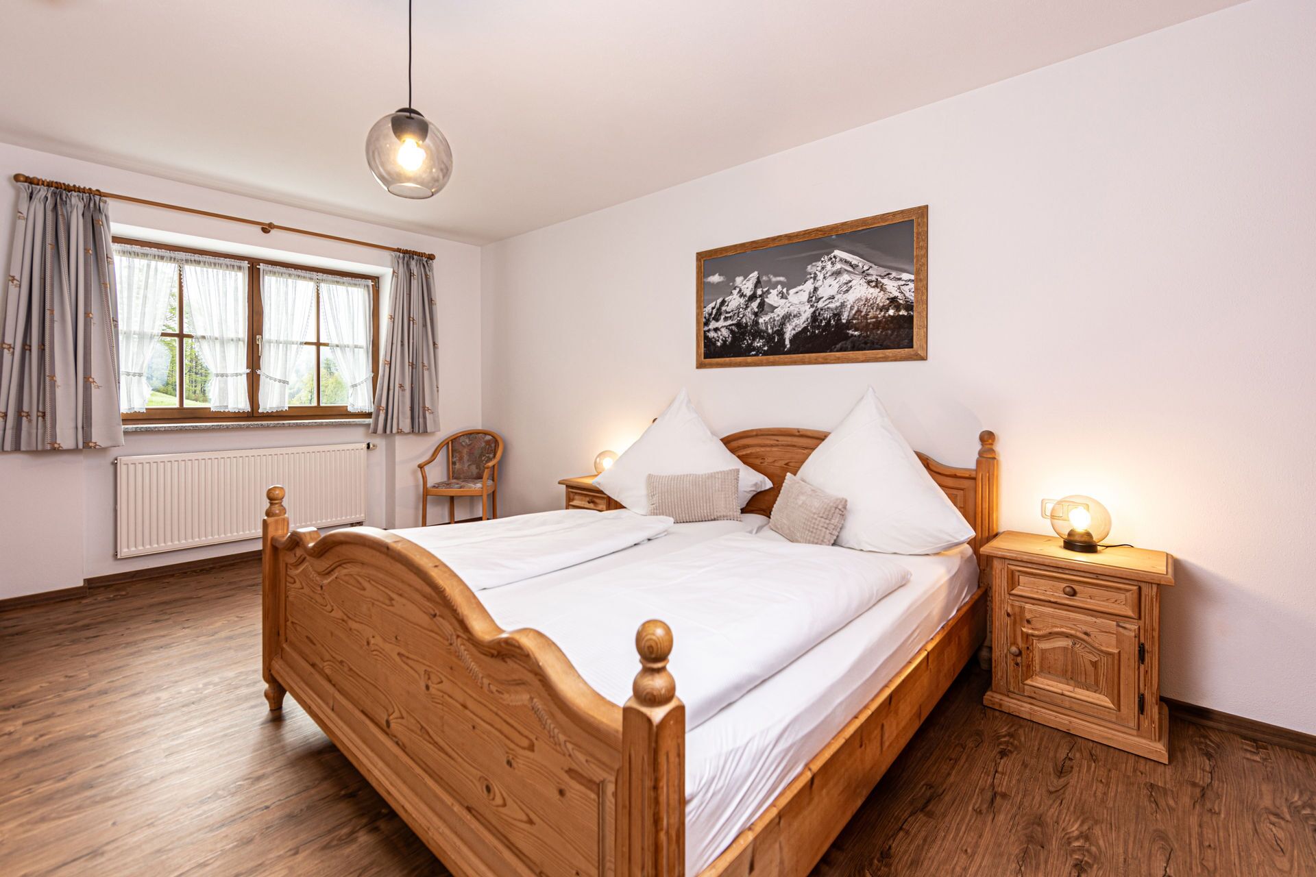 Quarto com cama de madeira, mesa de cabeceira com abajur, uma cadeira, janela com cortinas e pintura da paisagem de montanhas na parede.