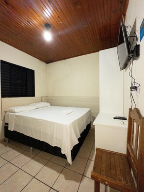 Una cama individual con sábana blanca, cabecero de madera, mesita de noche pequeña y televisor montado en la pared.