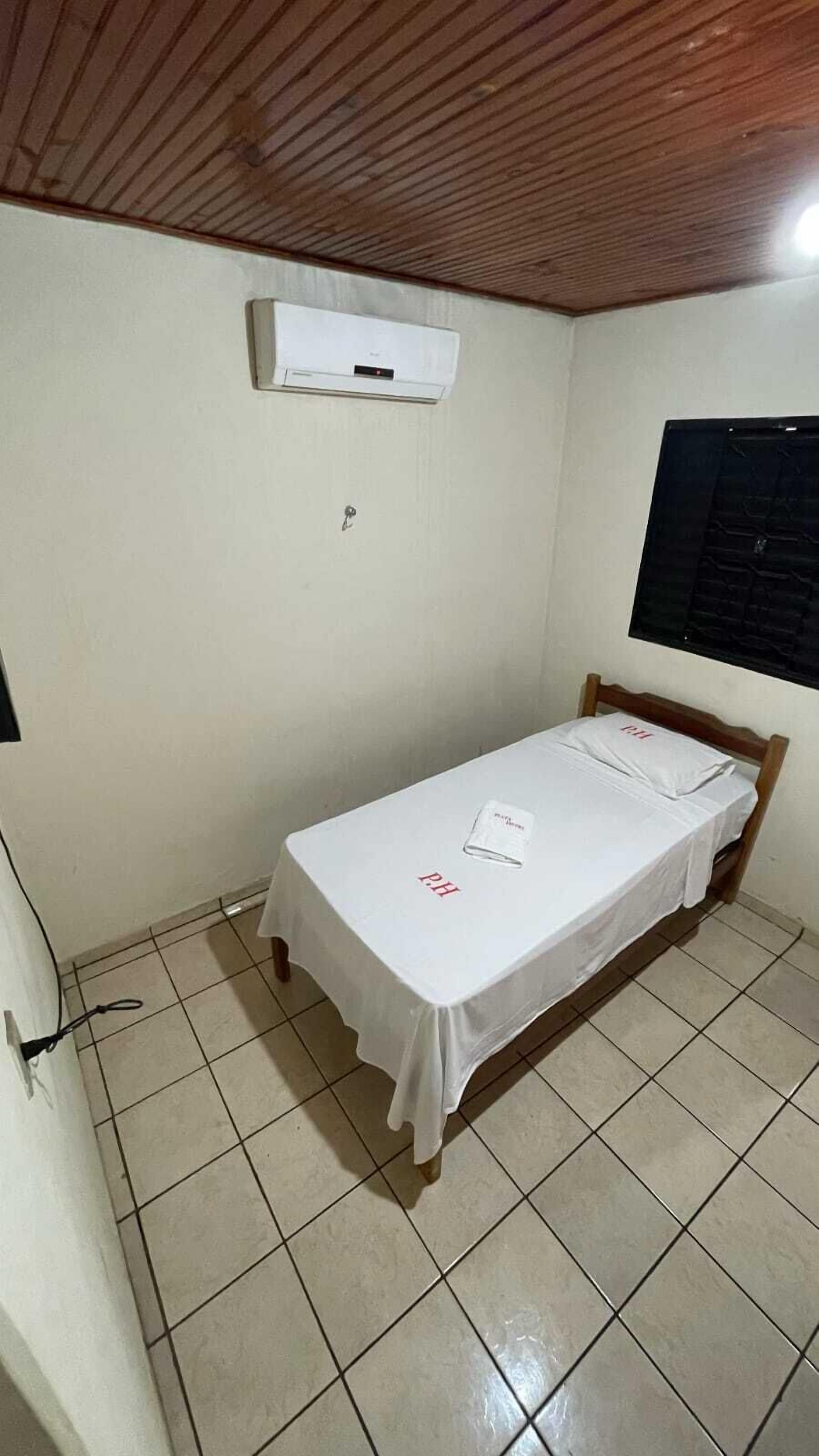 A small, single-bed room with a wooden ceiling, white walls, and a tiled floor. There is a window with a black blind, an air conditioning unit mounted on the wall, and a power outlet on the wall.