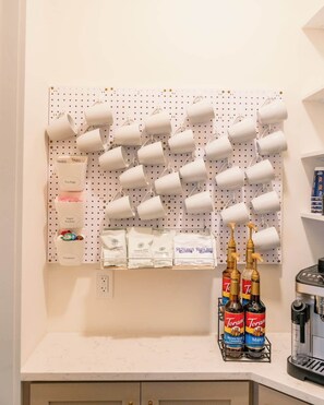 Una cocina con un panel de corcho que sostiene tazas y bolsitas de té, sobres de azúcar y cremas. También hay botellas de sirope Torani sobre la encimera.