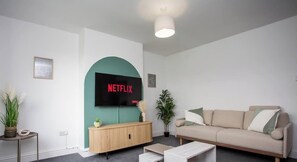 A living room with a TV mounted on a teal wall, a beige sofa, a wooden cabinet, and a small table with a plant.