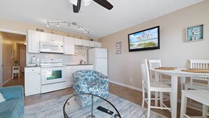 A modern kitchen with white cabinets, a microwave, and a stove. There is a dining area with a white table and chairs, and a blue patterned armchair. A ceiling fan and a flat-screen TV are visible. - Flash Sale. Direct Gulf Front 1/1 Sleeps 6 (Gulf Shores)