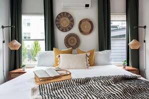 Ein ordentlich bezogenes Bett mit einer gestreiften Decke, zwei Nachttische mit Lampen und Blick durch große Fenster nach draußen.