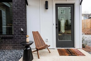 L’extérieur d’une maison moderne, avec un mur en briques sombres, une chaise en bois, une petite table et une porte dotée d’un panneau de verre.