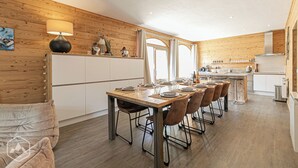 A modern kitchen with wooden paneling, a dining area with a long table and chairs, and a view of the outdoors through large windows.