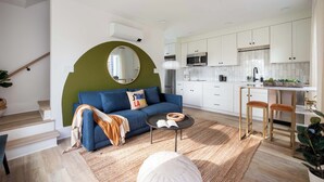 A modern living room with a blue sofa, a round mirror, and a small coffee table. A kitchen with white cabinets and a microwave is visible in the background.