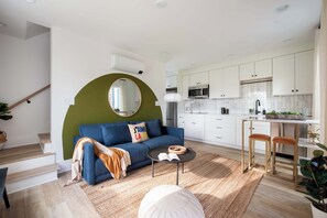 Un salon moderne avec un canapé bleu, un miroir rond et une petite table basse. On aperçoit, au fond, une cuisine avec des meubles blancs et un four à micro-ondes.