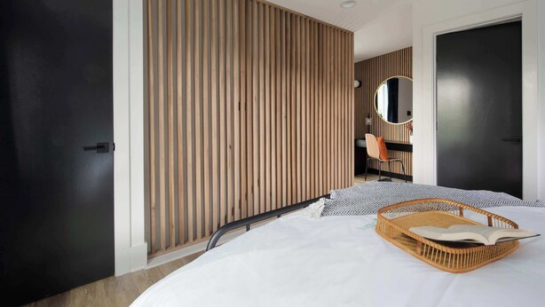 A modern bedroom with a bed, a wooden slatted wall, a round mirror, and a black door.