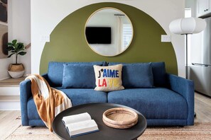 Uma sala de estar moderna com um sofá azul, um espelho redondo, uma televisão de tela plana e uma mesa de centro com um livro e uma cesta de palha.