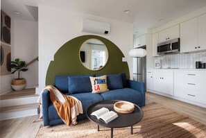 Una sala de estar moderna con un sofá azul, un espejo redondo, una mesa de centro con un libro y un tazón, y una zona de cocina con armarios blancos y un microondas.