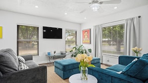 Ein Wohnzimmer mit grauem Sofa, blauem Sofa, rundem Couchtisch mit Blumenvase und Deckenventilator.