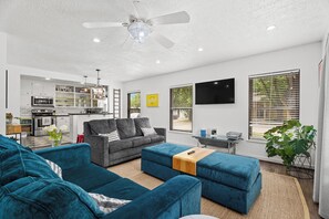 A modern living room with a grey sofa, blue ottomans, and a kitchen in the background. - Whiskey Ginger - Wine Barn, Fire Pit & More! (Fredericksburg)