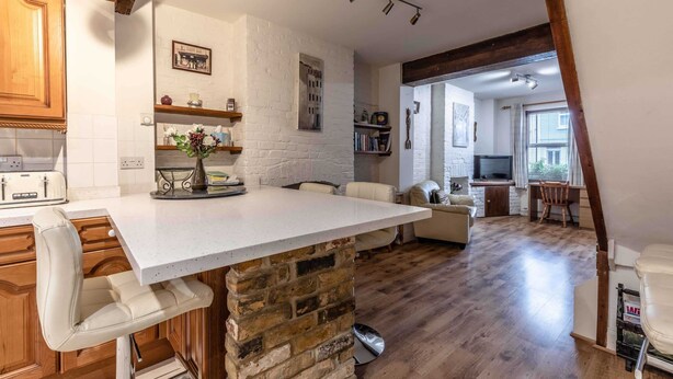 A kitchen with white brick walls, wooden cabinets, a white countertop, and a stone base. There is a dining area with a white sofa and a wooden table.