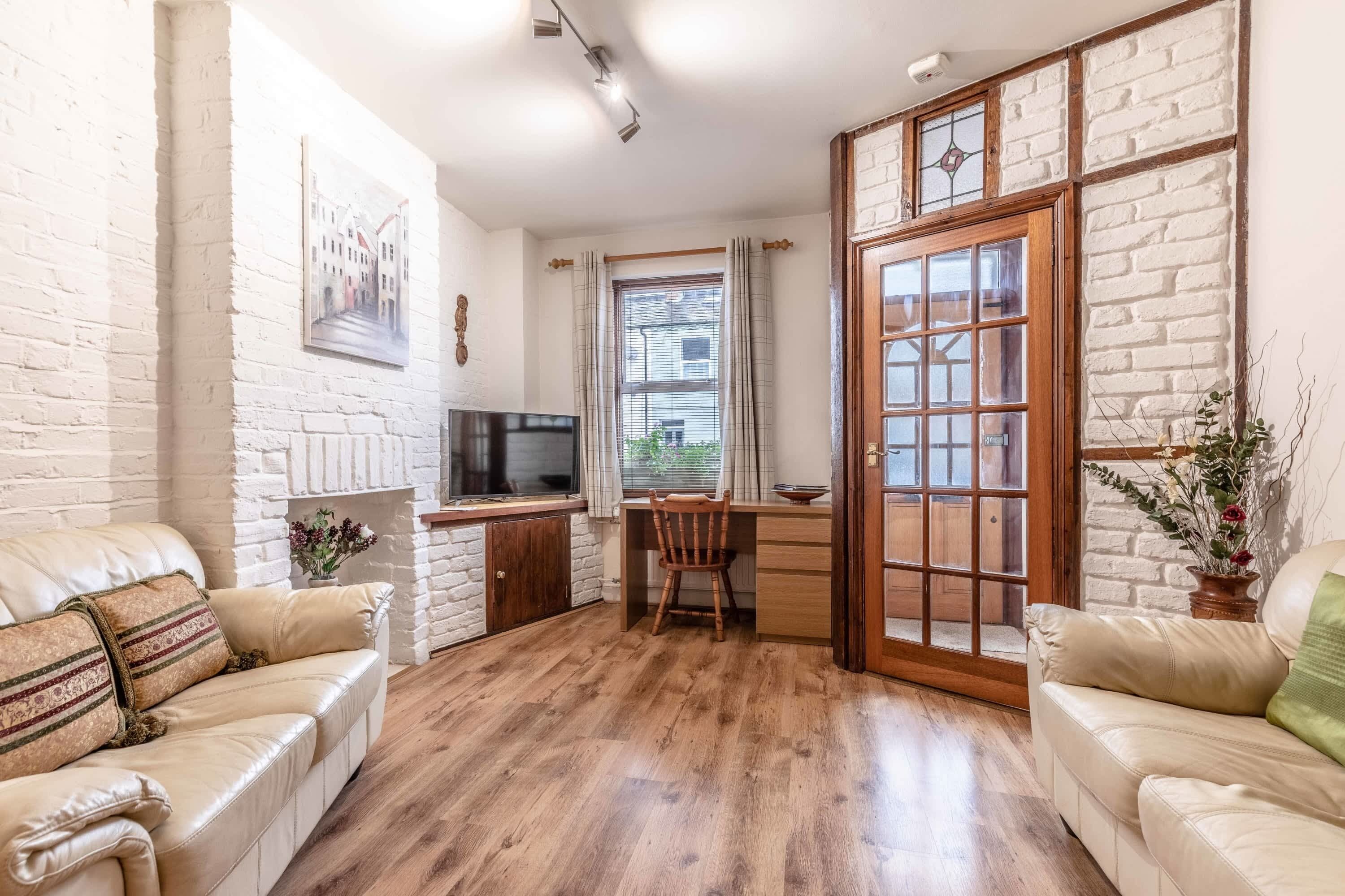 Una habitación con pared de ladrillo blanco, un escritorio de madera con un televisor, una silla de madera y dos sofás color beige.