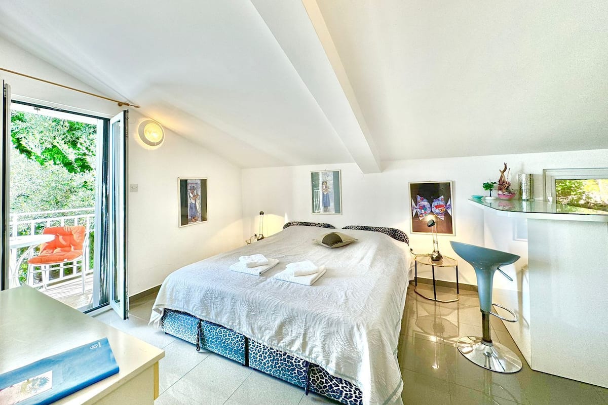A bedroom with a bed, a desk, a chair, and a balcony with a view of greenery.