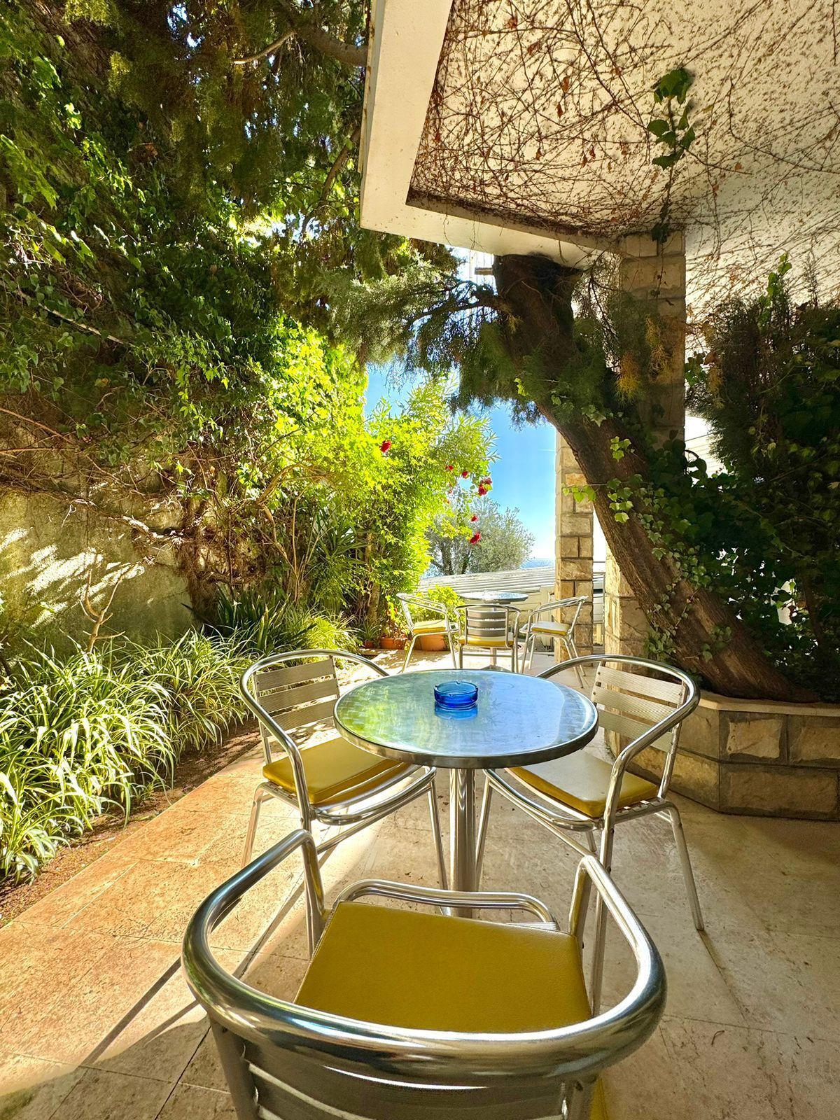 An outdoor patio with a round table and chairs, surrounded by greenery and a stone wall.
